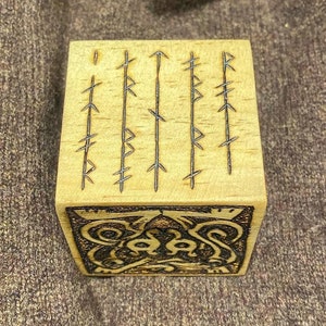 May include: A light-coloured wooden cube with etched runes and a decorative design. The cube features vertical lines of runes on the top and an intricate pattern on one side, showcasing a blend of craftsmanship and historical symbolism.