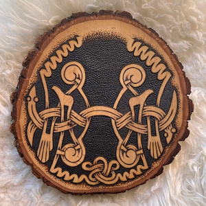May include: Round wooden plaque with a carved Viking design. The plaque features a light brown wood grain border and a dark, textured center. The design is a light brown, intricate knotwork pattern. The plaque is approximately 10 inches in diameter.
