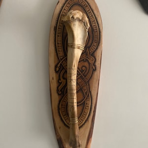 May include: A bone-shaped object, possibly a decorative piece, mounted on a wooden plaque. The plaque features intricate etched designs and runes, with a natural wood grain finish. Gold-colored wire wraps the bone.