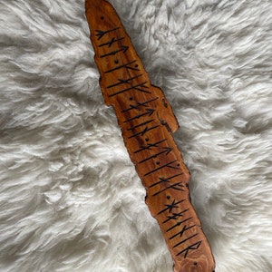 May include: A wooden rune stave with carved symbols. The stave is a natural brown color with dark brown etched runes. The wood has a rough, organic edge and is displayed on a white, fluffy surface.