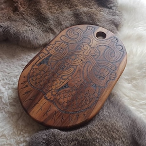 May include: A wooden, oval-shaped cutting board with intricate, carved Viking-style designs. The board is a warm brown color and features a hole for hanging. The carvings include a central figure and runic symbols along the edge.