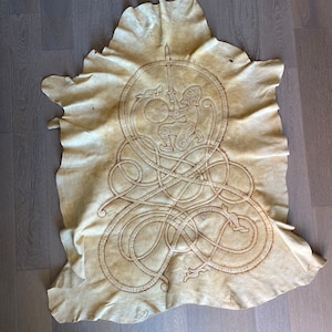 May include: A cream-colored leather hide featuring an embroidered Viking warrior design. The warrior is depicted with a sword and shield within a circular frame, surrounded by intricate knotwork. The hide has a natural, irregular shape.