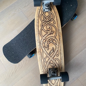 May include: Two skateboards. One is light wood with a detailed engraved design and black wheels. The other is black with black wheels. Both are on a light wood surface.