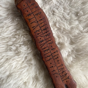 May include: A hand-carved wooden rune stave with dark etched symbols. The stave is a rich brown color and has a natural, textured surface. The runes are arranged in vertical lines with small dots and arrow-like symbols.