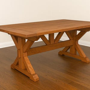 Może przedstawiać: Prostokątny drewniany stół jadalny z naturalnym wykończeniem. Stół ma podstawę w kształcie kozy z ukośnymi wspornikami i ma około 183 cm długości.