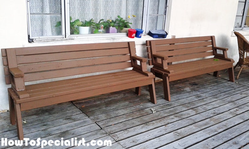 Pu&ograve; includere: Due panche in legno marrone con braccioli su un terrazzo in legno. Le panche hanno un design a doghe e sono dipinte in un caldo colore marrone. Lo sfondo comprende una finestra con piante e un muro bianco.
