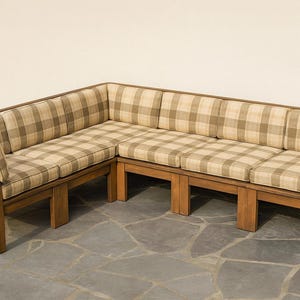 Puede incluir: Un sofá seccional en forma de L con estructura de madera y cojines a cuadros beige y marrón. El sofá tiene un perfil bajo y es adecuado para una sala de estar o zona de descanso. El marco de madera es de color marrón claro.