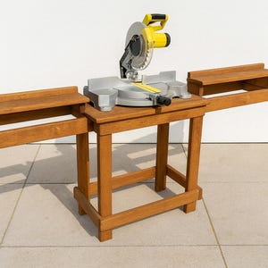 May include: A wooden miter saw stand with a yellow and silver miter saw. The stand has two side extensions and is made of light brown wood. The saw blade is visible. The stand is on a tiled floor.