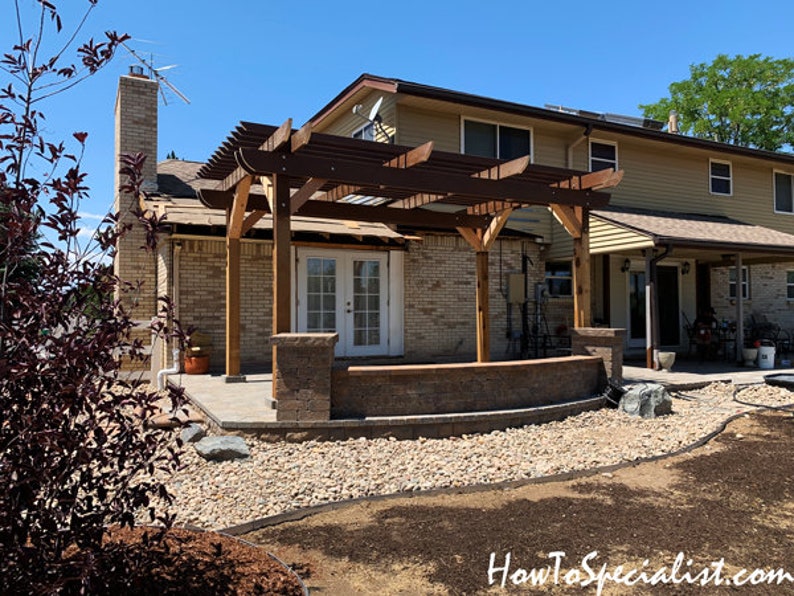 May include: A backyard patio with a wooden pergola, brick pillars, and a low brick wall. The patio area features a stone pathway and a landscaped yard with a house in the background. The sky is clear and blue.