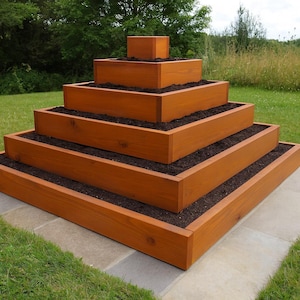 Puede incluir: Una jardinera de madera escalonada con cinco niveles, llena de tierra oscura. La jardinera está teñida de un marrón cálido y se asienta sobre un patio de piedra, rodeada de hierba verde y árboles. La estructura mide unos 152 cm de ancho.