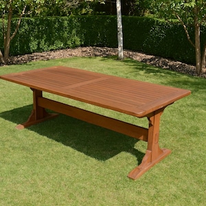May include: A rectangular brown wooden outdoor table with a slatted top and trestle-style legs. The table is set on a green lawn, surrounded by trees and bushes. The table measures approximately 72 inches long.