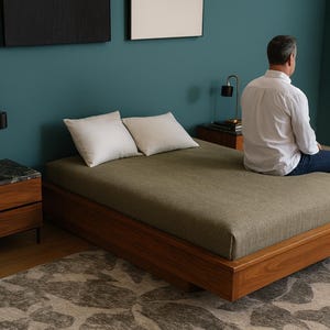 May include: A modern bedroom scene featuring a low-profile wooden bed with a green mattress and two white pillows. The room includes two wooden nightstands with lamps, a patterned rug, and artwork on the teal wall.