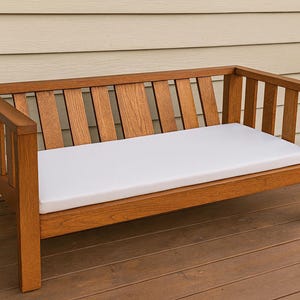Puede incluir: Banco de madera con cojín blanco. El banco tiene respaldo de listones y reposabrazos. La madera tiene un tono cálido, rojizo-marrón. El cojín es blanco y parece cómodo.
