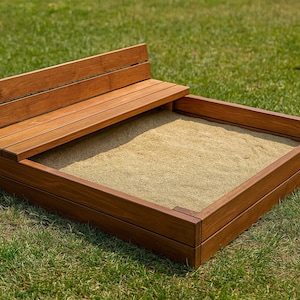 May include: A wooden sandbox with a built-in bench. The sandbox is rectangular with a dark brown finish, filled with sand. The bench is attached to the side and is made of the same wood. The sandbox sits on a bed of green grass.