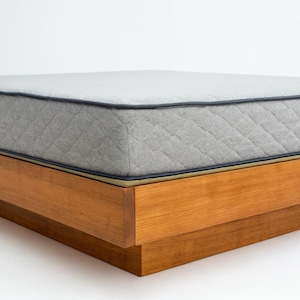 Może przedstawiać: Nowoczesne, niskoprofilowe łóżko z jasnoszarą matą. Materac ma pikowany wzór i jest obszyty ciemnoniebieską lamówką. Rama łóżka wykonana jest z jasnobrązowego drewna.