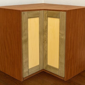 Może przedstawiać: Drewniana szafka narożna z czerwonobrązowym wykończeniem. Szafka posiada dwoje drzwi z jasnym panelem drewnianym obramowanym ciemniejszym drewnem. Szafka jest przeznaczona do wpasowania w róg.