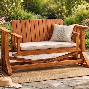 Puede incluir: Un banco columpio de madera para exteriores con un cojín beige claro y una almohada decorativa con un diseño de helecho. El banco está hecho de madera natural y está colocado sobre una alfombra tejida en un jardín.