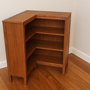 May include: A wooden corner bookshelf with a warm brown finish. The bookshelf has three shelves and is designed to fit into a corner. The wood grain is visible. The bookshelf is on a hardwood floor.