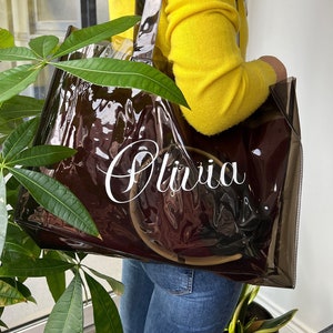 May include: Clear plastic tote bag with a brown lining and the name "Olivia" printed in white on the front.