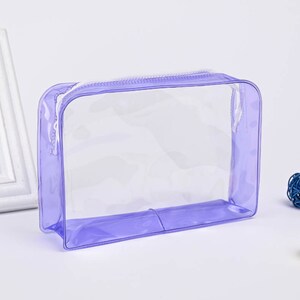 May include: A clear, lavender-colored zippered pouch with a white zipper. The pouch is made of a flexible, waterproof material.