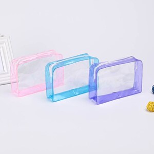 May include: Three clear plastic zippered pouches in pink, blue, and purple. The pouches are lying flat on a white surface.