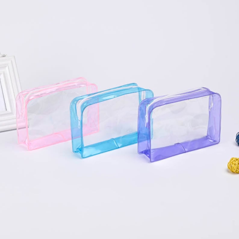 May include: Three clear plastic zippered pouches in pink, blue, and purple. The pouches are lying flat on a white surface.