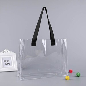 Peut inclure: Un sac fourre-tout transparent en plastique avec des poignées noires. Le sac est vide et posé sur une surface grise.