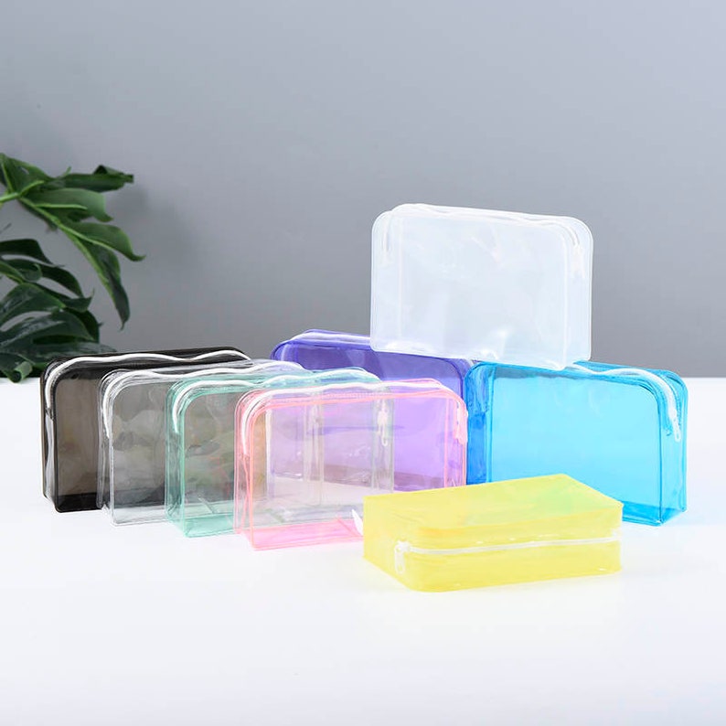 May include: Set of eight clear plastic zippered pouches in various colors, including black, green, pink, purple, blue, and yellow. The pouches are all rectangular and have a zipper closure.