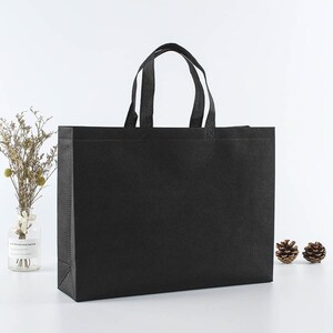 May include: A black non-woven tote bag with two black handles. The bag is sitting on a white surface.