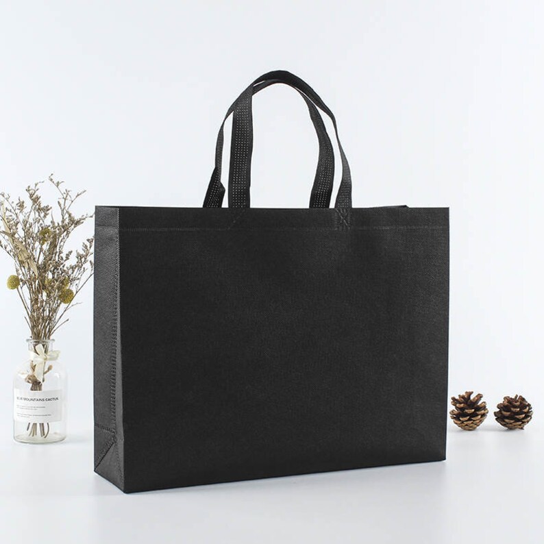 May include: A black non-woven tote bag with two black handles. The bag is sitting on a white surface.