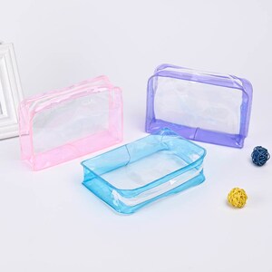 May include: Three clear plastic zippered pouches in pink, blue, and purple. The pouches are empty and are sitting on a white surface.