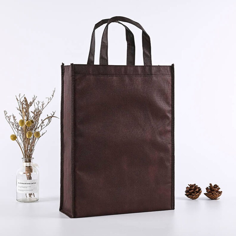 May include: A brown reusable tote bag with black handles. The bag is made of a non-woven fabric and has a smooth, matte finish.