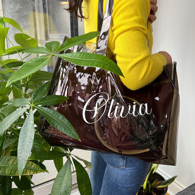 May include: Clear plastic tote bag with a brown lining and the name "Olivia" printed in white on the front.