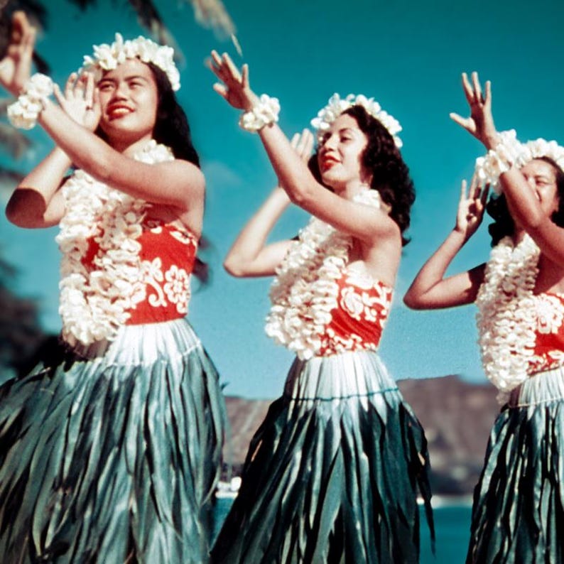 May include: Three women in traditional Hawaiian attire perform a dance. They wear red and white tops, long green skirts, and white flower leis and wristlets. The background is a clear blue sky.
