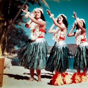 May include: Three women in traditional Hawaiian attire perform a hula dance on a sandy beach. They wear red and white tops, green skirts, and flower leis. The background includes palm trees and a blue sky.