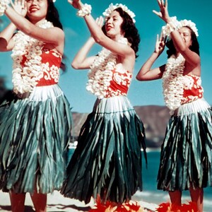 May include: Three women in traditional Hawaiian attire perform a dance. They wear red and white tops, green skirts, and white flower leis. The background shows a beach and blue sky, creating a vibrant scene.