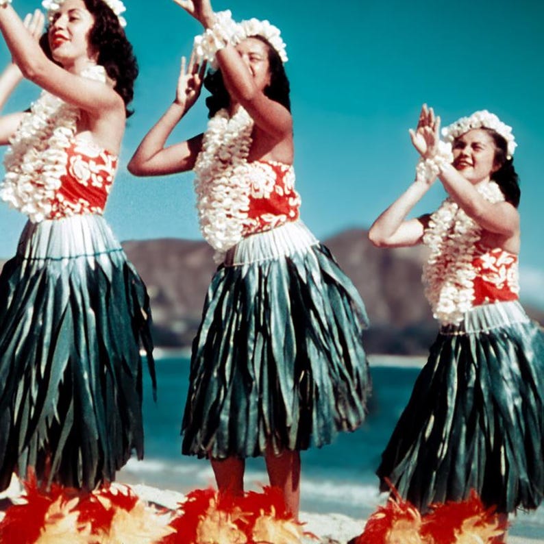 May include: Three women in traditional Hawaiian attire perform a dance on a beach. They wear white flower leis, red and white tops, and dark green skirts. Red and yellow feather accents adorn their feet, with a blue sky and ocean in the background.