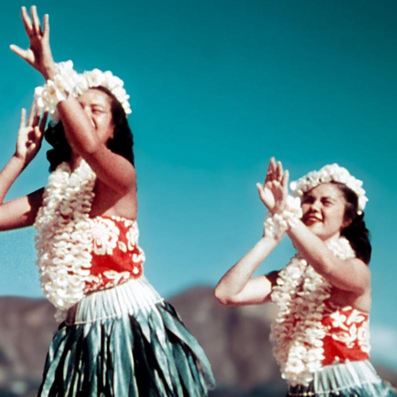 May include: Two women in traditional Hawaiian attire dance outdoors. They wear white flower leis, red and white tops, and skirts with blue and green accents. The background is a clear blue sky.
