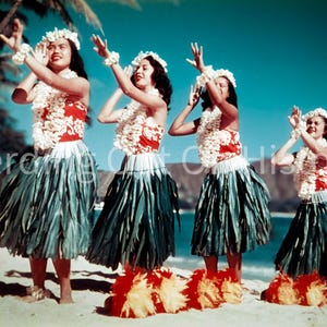 May include: Four women in traditional Hawaiian attire perform a dance on a sandy beach. They wear floral leis, headbands, and wristbands, along with red and white tops, green skirts, and feathered ankle decorations. The background features a blue sky and ocean.
