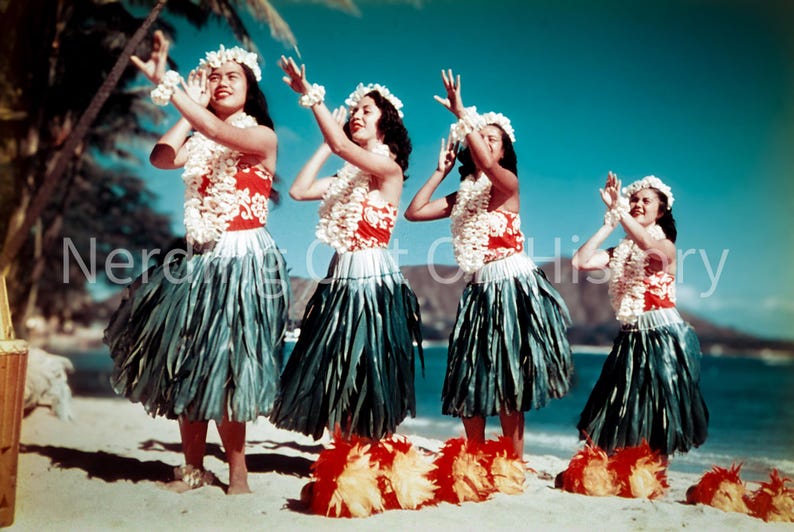 May include: Four women in traditional Hawaiian attire perform a dance on a sandy beach. They wear floral leis, headbands, and wristbands, along with red and white tops, green skirts, and feathered ankle decorations. The background features a blue sky and ocean.