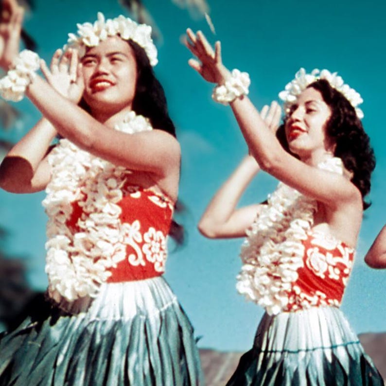 May include: Two women in traditional Hawaiian attire perform a dance. They wear red and white floral tops, long green skirts, and white flower leis and headbands. The background is a bright blue sky.
