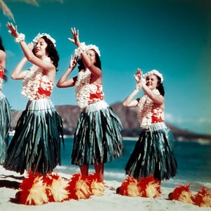 May include: A vintage photograph captures a group of women performing a traditional Hawaiian dance on a sandy beach. They wear floral leis, headbands, and skirts made of green leaves. Their tops are red with white floral patterns, and their feet are adorned with red and yellow accents.