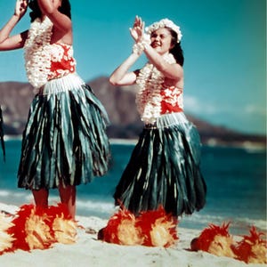 May include: Two women in traditional Hawaiian attire perform a hula dance on a sandy beach. They wear red and white tops, blue skirts, and floral leis. The scene is set against a backdrop of the ocean and a clear blue sky.