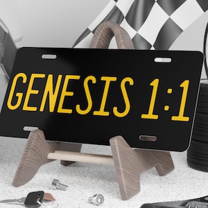 May include: Black license plate with the text "GENESIS 1:1" in yellow, displayed on a wooden stand. A racing flag, tires, helmet, and tools are in the background, suggesting a racing or automotive theme.
