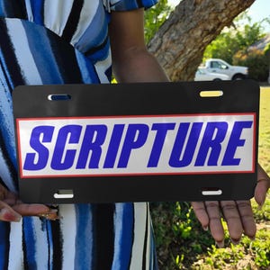 May include: Black license plate with the word "SCRIPTURE" in large, blue, bold letters. The text is set against a white background with a red border. The license plate is held up in front of a person wearing a blue and white striped dress.