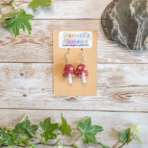 May include: Handmade crochet mushroom earrings. The earrings have red caps with white spots and white stems. They are displayed on a brown card with the text "Harriet's Macrame". The background is a light wood surface.