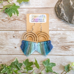 May include: A pair of rainbow-colored macrame earrings with wooden rainbow arches. The earrings are handmade with recycled cotton and are labeled "Harriet's Macrame".