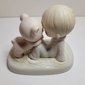 Może przedstawiać: Porcelanowa figurka przedstawiająca chłopca i prosię, które się tulą. Chłopiec ma na sobie białą i brązową koszulę, a prosię jest różowe. Figurka stoi na białej podstawie.