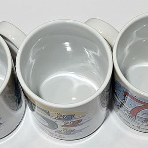 May include: Six white ceramic mugs with colorful designs. Each mug features a different number, from 1 to 6, and a unique illustration. The mugs are arranged in a row, with the numbers increasing from left to right.