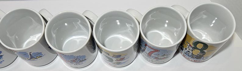 May include: Six white ceramic mugs with colorful designs. Each mug features a different number, from 1 to 6, and a unique illustration. The mugs are arranged in a row, with the numbers increasing from left to right.